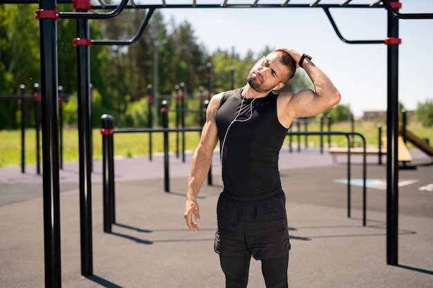 Jeune homme musclé en vêtements de sport et écouteurs faisant de l'exercice pour le cou en se tenant debout sur le terrain de sport