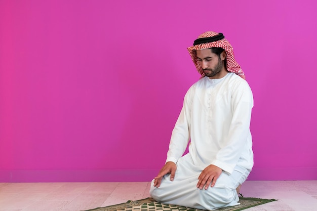 Jeune homme musulman arabe en vêtements traditionnels priant sur le tapis devant le mur rose avant le dîner de l'iftar lors d'une fête du ramadan à la maison.