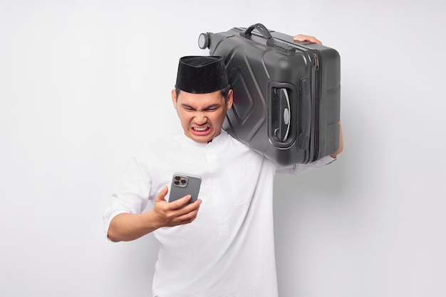 Jeune homme musulman asiatique en colère portant une valise et utilisant un téléphone portable tout en réagissant aux mauvaises nouvelles isolées sur fond blanc Concept de Ramadan et eid Mubarak