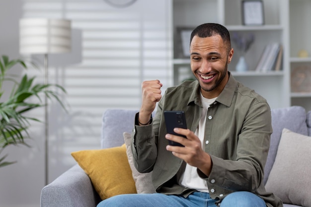 Jeune homme musulman souriant utilisant un téléphone assis à la maison sur un canapé jouant à des jeux en ligne plaçant des paris