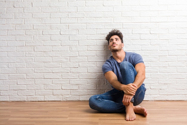 Jeune homme naturel assis sur un plancher en bois en levant, pensant à quelque chose d&#39;amusant et ayant un