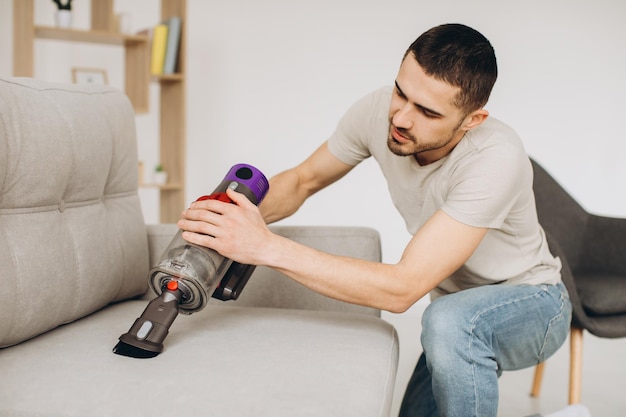 Un jeune homme nettoie le canapé avec un aspirateur