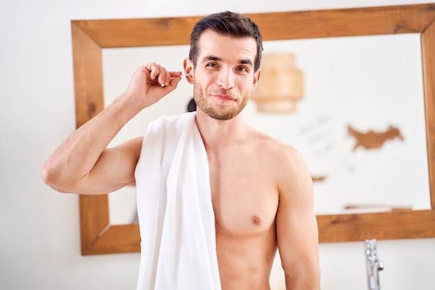 Un jeune homme nettoie son oreille avec un coton-tige en se tenant debout dans le miroir en face du bain le matin