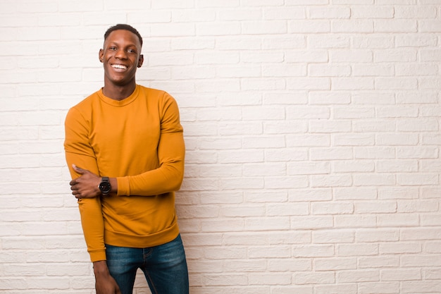 Jeune homme noir afro-américain rire timidement et joyeusement, avec une attitude amicale et positive mais peu sûre contre le mur de briques