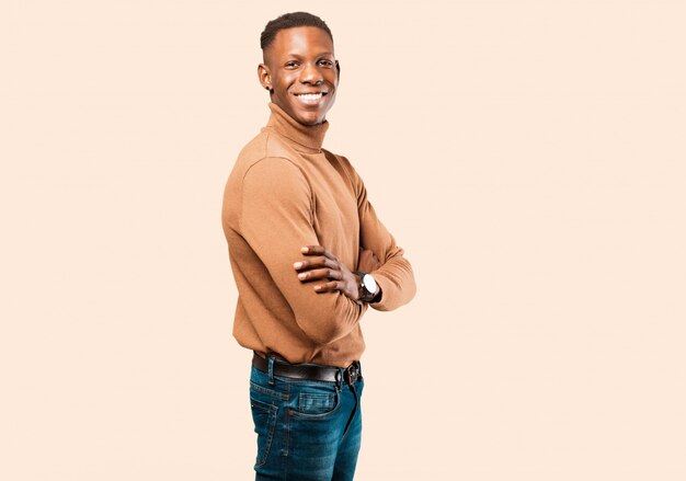 Jeune homme noir afro-américain souriant à la caméra avec les bras croisés et une expression heureuse, confiante et satisfaite, vue latérale contre le mur beige