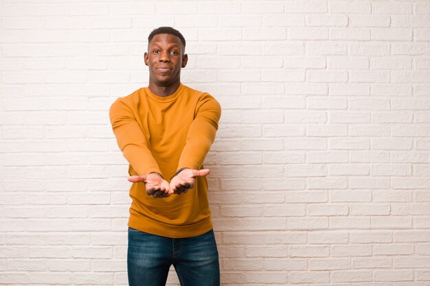 Jeune homme noir afro-américain souriant joyeusement avec un regard amical, confiant et positif, offrant et montrant un objet ou un concept contre le mur de briques