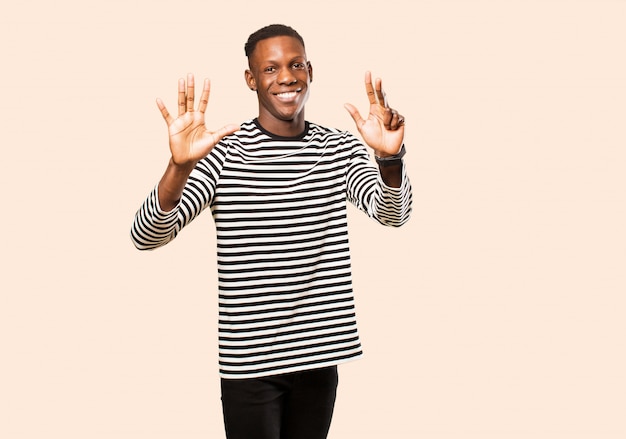 Jeune homme noir afro-américain souriant et regardant amical, montrant le numéro sept ou septième avec la main vers l'avant, compte à rebours contre le mur beige