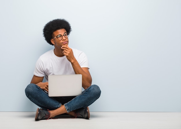 Jeune homme noir assis sur le sol avec un ordinateur portable doutant et confus