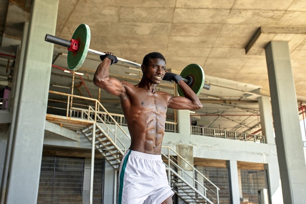 Jeune homme noir athlétique soulevant une barre de poids lourd dans une salle de sport en plein air sous le pont Concept de mode de vie sain