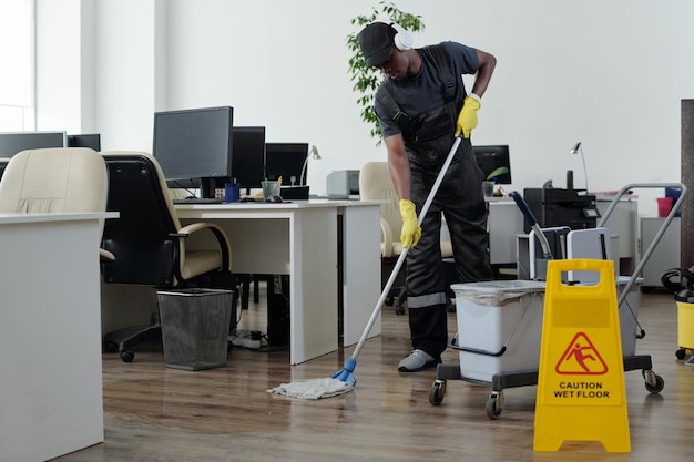 Jeune homme noir contemporain en vêtements de travail nettoyant le sol dans un bureau à espace ouvert