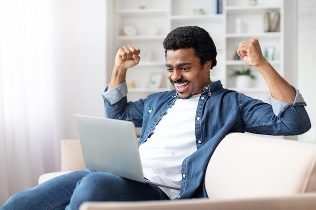 Un jeune homme noir excité célèbre son succès à la maison avec un ordinateur portable.