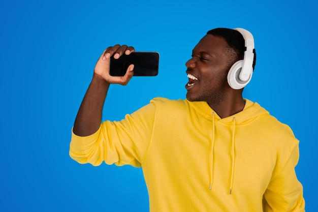 Un jeune homme noir heureux dans des écouteurs sans fil chante sur un smartphone avec un écran vide dans l'imaginaire