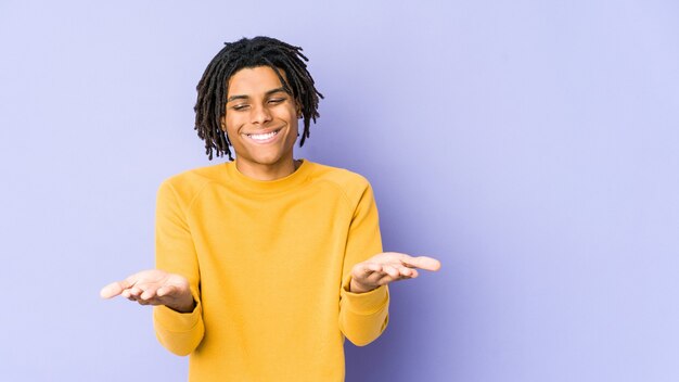 Photo jeune homme noir portant une coiffure rasta tenant quelque chose avec des paumes, offrant à la caméra.