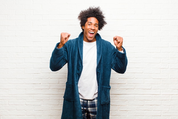 Jeune homme noir portant un pyjama avec une robe se sentant heureux, surpris et fier, criant et célébrant le succès avec un grand sourire sur le mur de briques