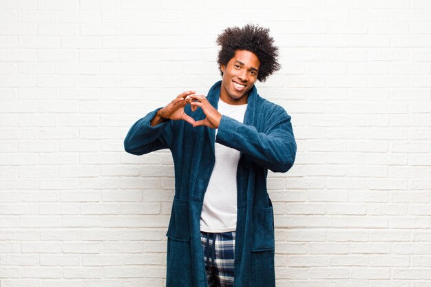 Jeune homme noir portant un pyjama avec une robe souriante et se sentant heureux, mignon, romantique et amoureux, faisant forme de coeur avec les deux mains contre le mur de briques