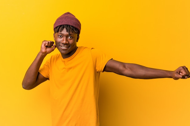 Jeune homme noir portant des rastas sur un mur jaune, dansant et s'amusant.