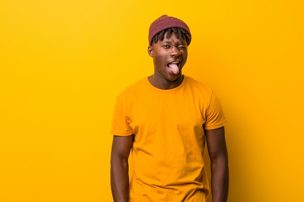 Jeune homme noir portant des rastas sur le mur jaune drôle et sympathique qui lui sort la langue.