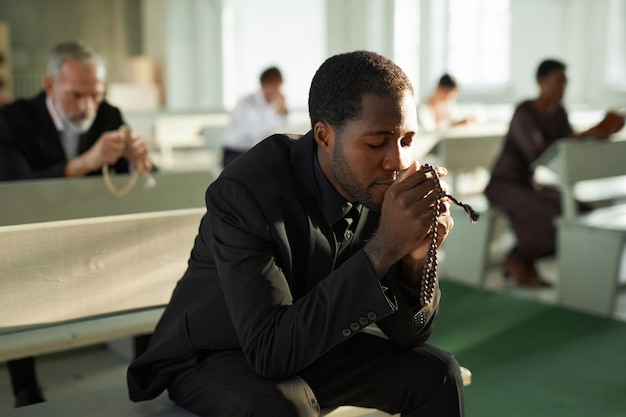 Un jeune homme noir en prière silencieuse à l'église tenant un chapelet à la lumière du soleil