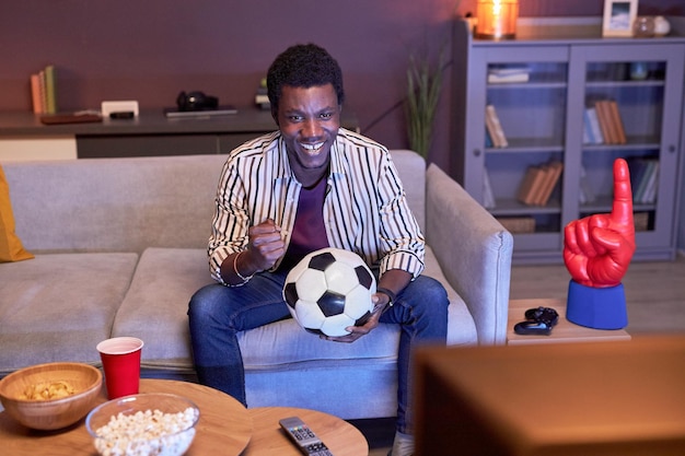 Jeune homme noir regardant un match de football à la télévision à la maison et tenant le ballon