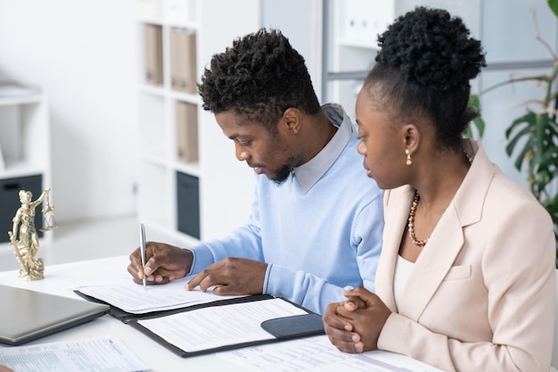 Jeune homme noir sérieux assis à table dans un bureau d'avocats et signant des papiers de divorce