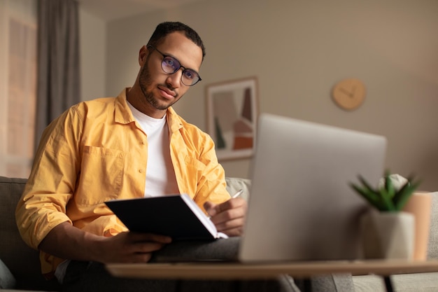 Jeune homme noir sérieux utilisant un ordinateur portable travaillant à distance en prenant des notes lors d'une réunion d'affaires à distance à