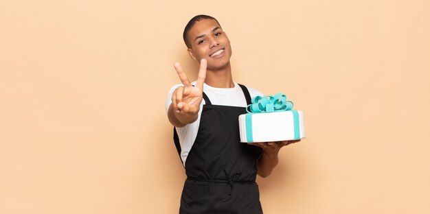 Jeune homme noir souriant et regardant heureux, insouciant et positif, faisant des gestes de victoire ou de paix d'une main