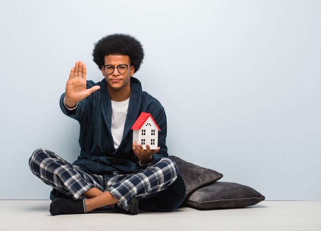 Jeune homme noir tenant un modèle de maison assis sur le sol en mettant la main devant