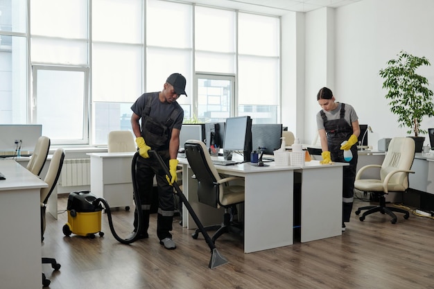 Photo jeune homme noir en vêtements de travail nettoyant le sol pendant qu'une fille essuyait des bureaux avec des ordinateurs