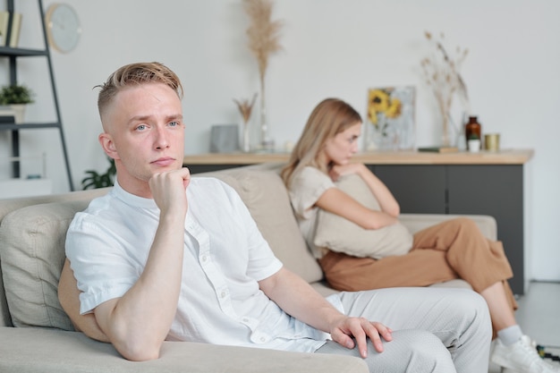 Jeune homme offensé assis sur un canapé avec sa femme malheureuse en arrière-plan tout en s'attendant à ce qu'elle parle d'abord après une dispute