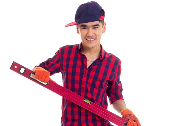 Jeune homme optimiste en chemise à carreaux rouge avec des gants orange et un snapback bleu tenant le niveau rouge