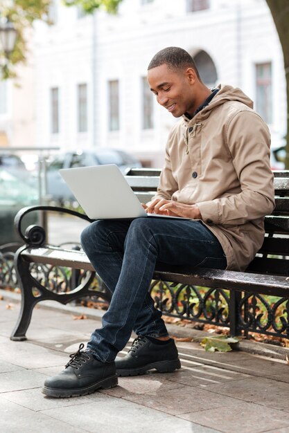 Jeune homme avec ordinateur portable assis sur un banc en bois et en tapant