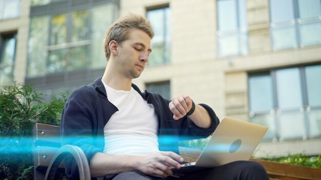 Jeune homme avec ordinateur portable consultant sa montre fond de lumière parasite maison