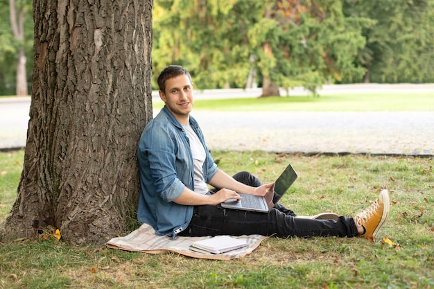 Jeune homme avec ordinateur portable reste dans l'herbe sur le campus universitaire.