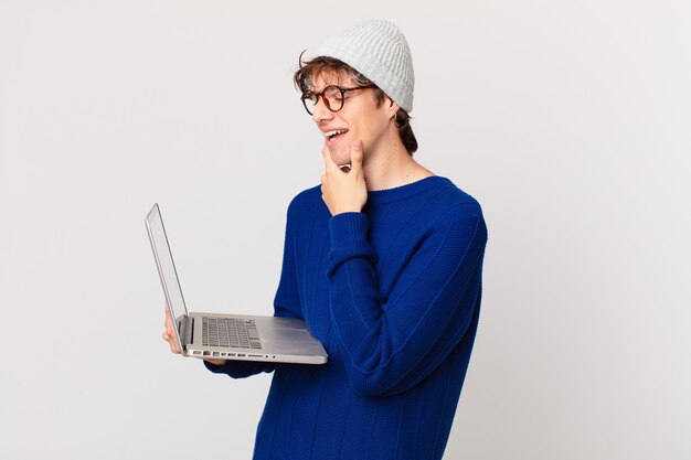 Jeune homme avec un ordinateur portable souriant avec une expression heureuse et confiante avec la main sur le menton