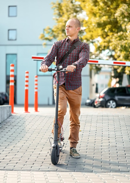 Jeune homme en pantalon décontracté et chemise debout à côté de son scooter électrique, regardant de côté