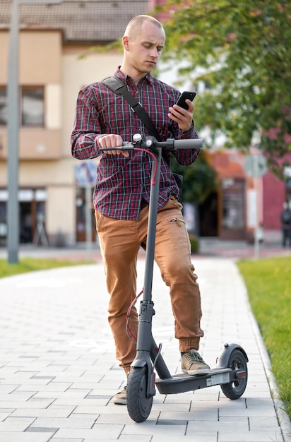 Jeune homme en pantalon décontracté et chemise debout à côté de son scooter électrique, vérifiant le téléphone portable