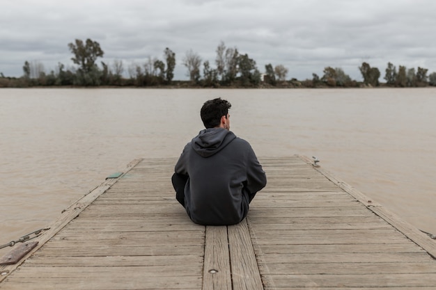 Jeune homme par derrière assis dans une jetée à côté d'une rivière tout en regardant et se reposant avec un ciel nuageux