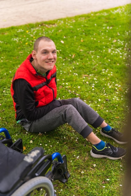 Un jeune homme paralysé assis sur l'herbe à côté du fauteuil roulant à côté d'un arbre regardant la caméra