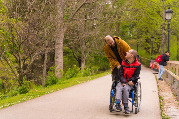 Photo jeune homme paralysé en fauteuil roulant poussé par un ami dans un parc public de la ville se promenant le long d'un chemin s'amusant au printemps