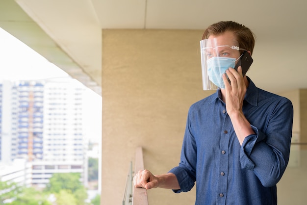 Jeune homme parlant au téléphone avec masque et écran facial sur la vue de la ville