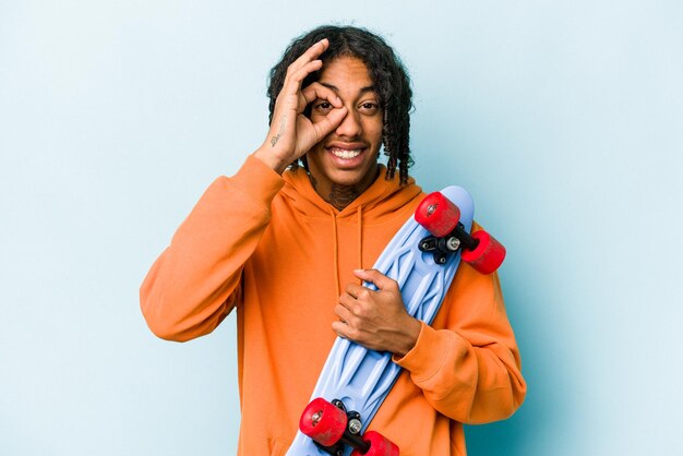 Jeune homme patineur afro-américain isolé sur fond bleu excité en gardant le geste ok sur les yeux