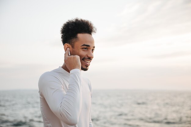 Un Jeune Homme à La Peau Sombre Et Frisée, En T-shirt Blanc, Sourit Sincèrement Et écoute De La Musique Dans Des écouteurs Sans Fil Près De La Mer