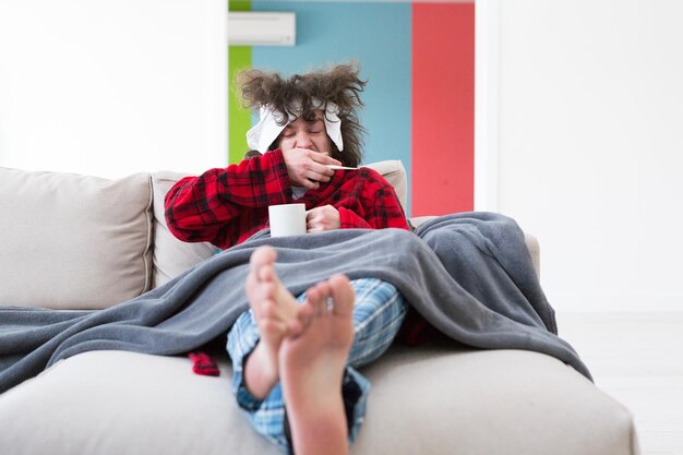 Photo jeune homme en peignoir avec grippe et fièvre enveloppé tenant une tasse de thé de guérison assis sur un canapé à la maison