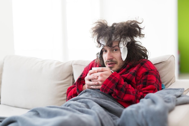Photo jeune homme en peignoir avec grippe et fièvre enveloppé tenant une tasse de thé de guérison assis sur un canapé à la maison