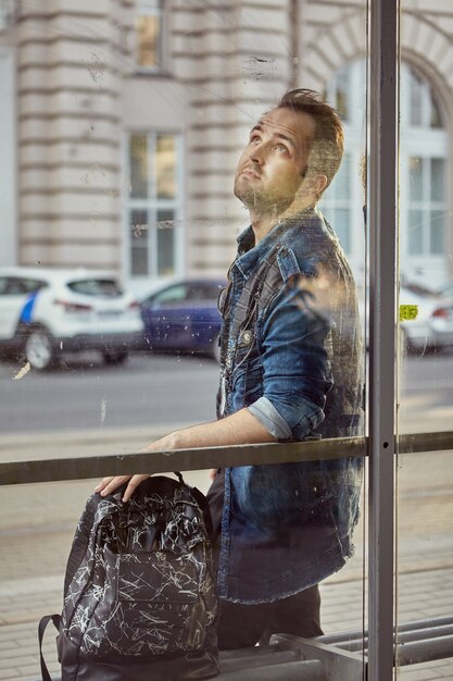 Jeune homme pensif est debout sur la station de transport public avec sac à dos.