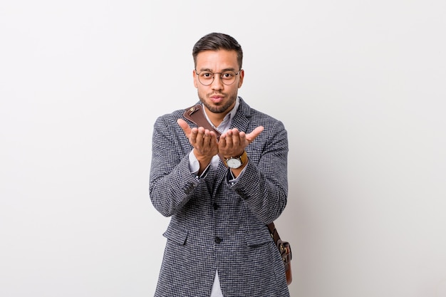 Jeune homme philippin d'affaires contre un mur blanc se pliant les lèvres et tenant les paumes pour envoyer l'air baiser.