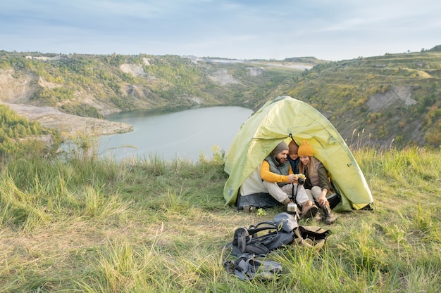 Jeune homme avec photocaméra montrant des photos à son petit fils et à sa femme assis près d'une tente après le dîner dans un environnement naturel le week-end