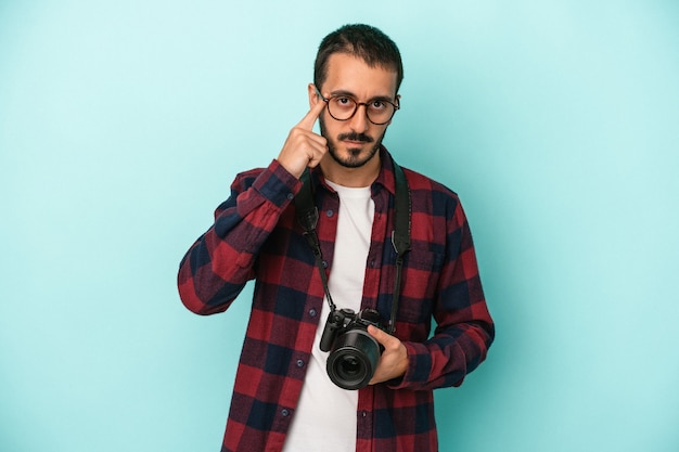 Jeune homme photographe caucasien isolé sur fond bleu pointant le temple avec le doigt, pensant, concentré sur une tâche.