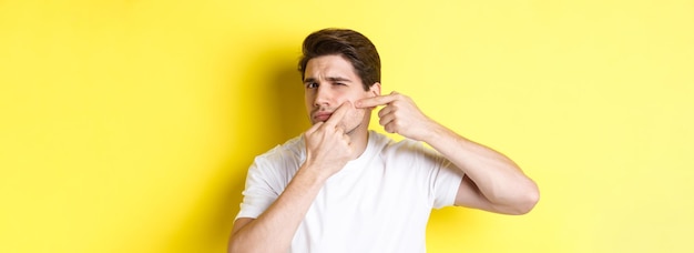 Jeune homme pop un bouton sur la joue debout sur fond jaune concept de soins de la peau et de l'acné