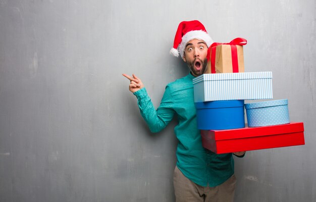 Jeune homme portant un bonnet de Noel tenant des cadeaux pointant vers le côté